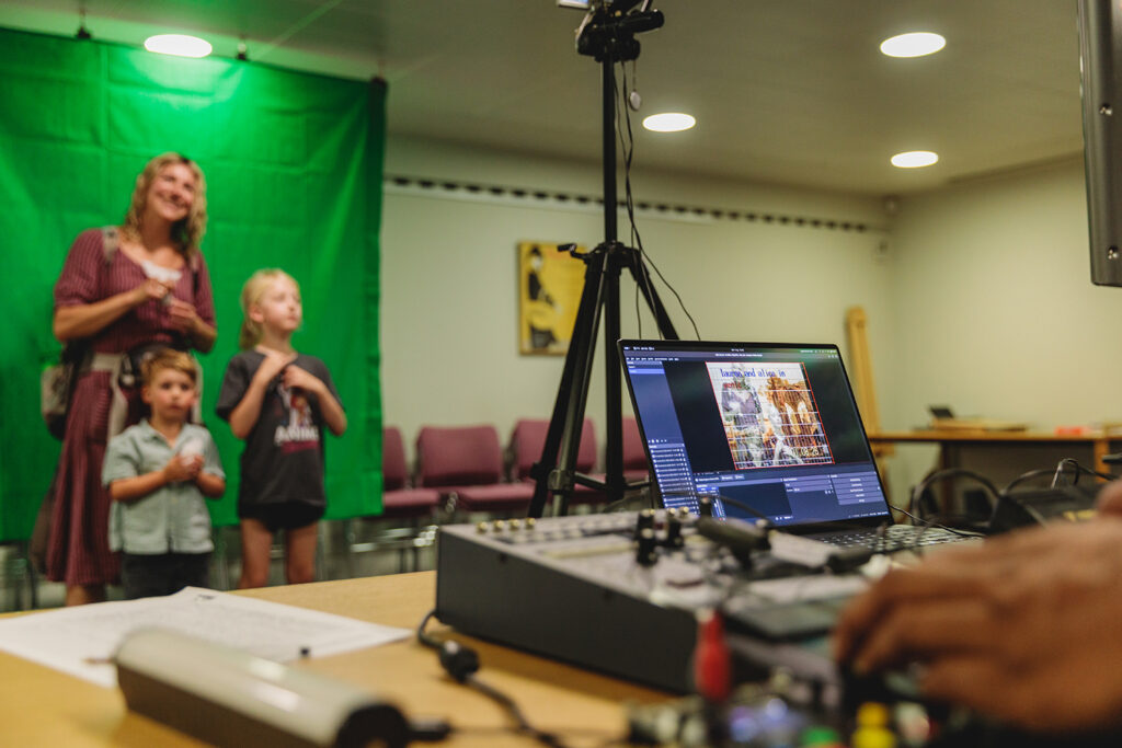A family, one woman and two small children, stand in front of a green screen in a room at the Fitzwilliam Museum. They appear blurred in the background while in the foreground we see a laptop and editing software on a wooden table.