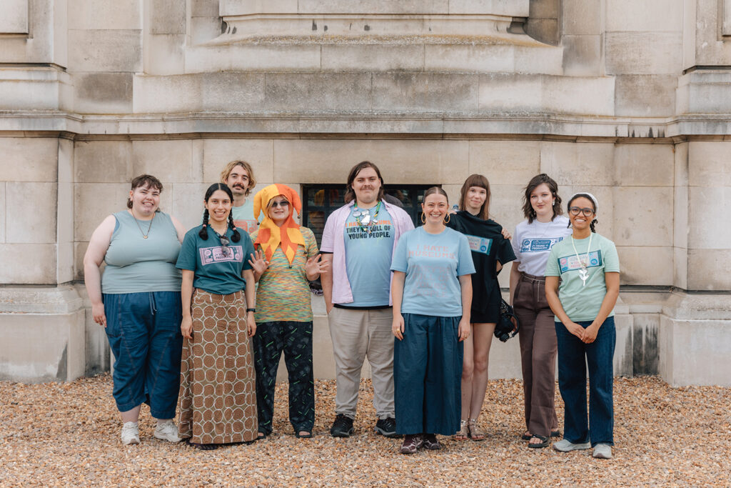 The Young Producers, a group of nine young people, stand smiling in a line outside the stone exterior of the Fitzwilliam Museum.