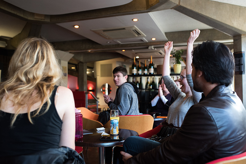 Players seated in a bar. One girl is smiling with her hands in the air in a celebratory gesture