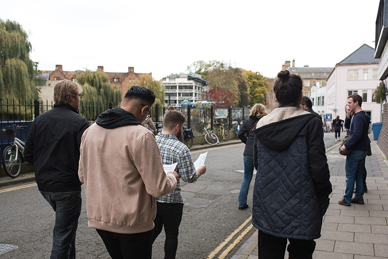 Group of four players shown from the back as they walk down a lane by the river, looking at their maps