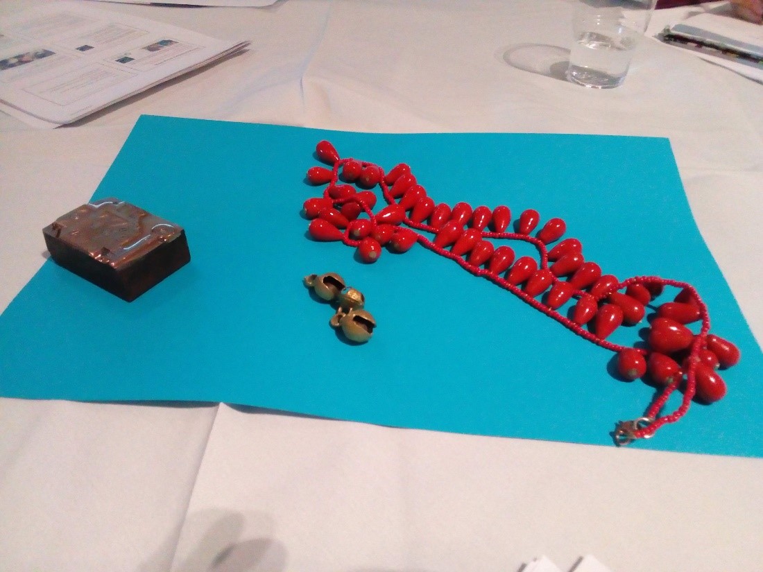 A selection of three mystery objects laid out on blue paper: a shiny string of red beads on a red cord; three small round metal objects; a small black box with a patterned lid