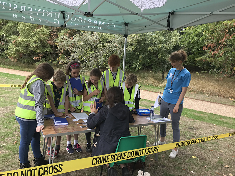 A cluster of children and a museum educator gather around a table under an awning on a lawn in the Botanic Garden. Yellow plastic tape extends across the picture saying "Crime Scene Do Not Enter"