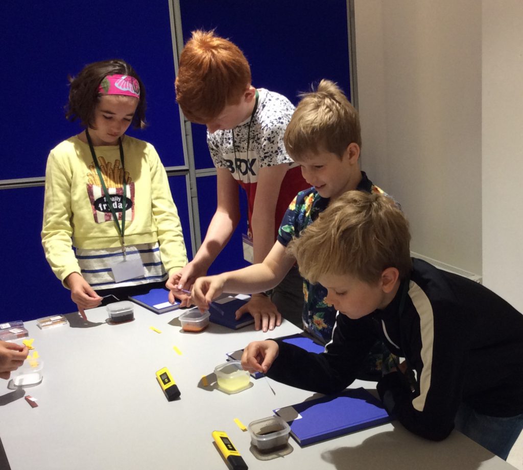 Four children use litmus papers to test the pH of different coloured soil samples in small plastic tubs