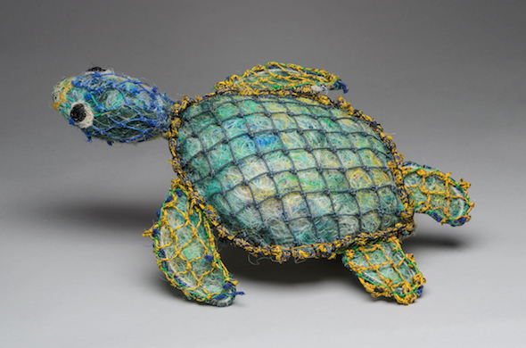 A sculpture of a turtle made from blue plastic fibres wrapped in recycled yellow and blue netting, displayed against a grey background as if swimming towards the edge of the frame.