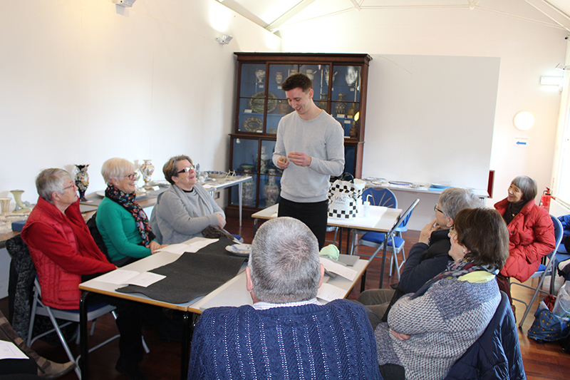 A group of smiling volunteers sit around a table listening to Tim, who is standing at one end