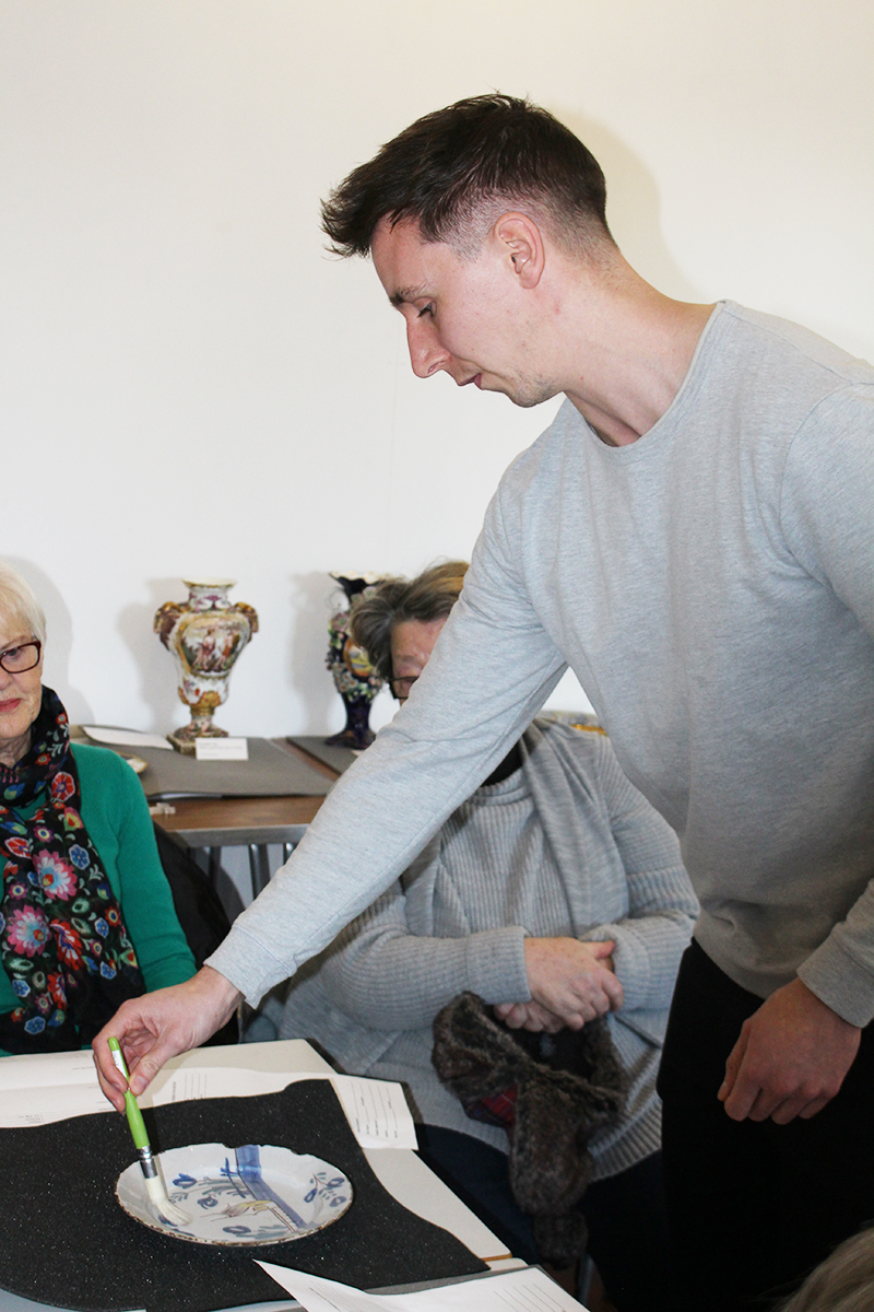 Tim delicately brushes a small blue and white plate as two volunteers look on