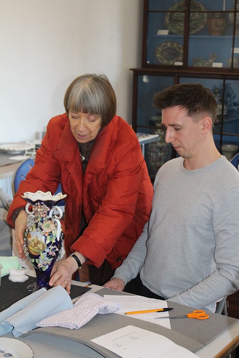 Julia places a large, elaborate blue and white vase on the table in front of Tim