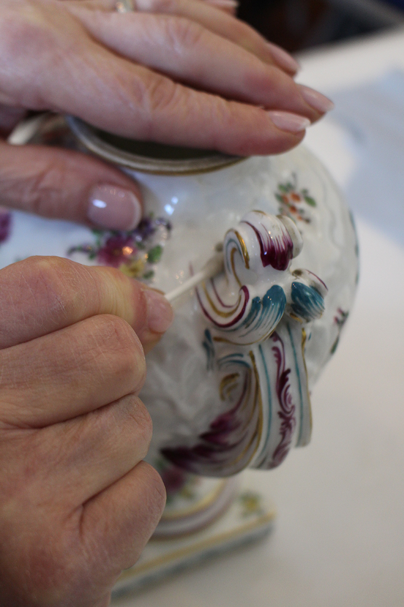 A close up of two hands, one stabilising a white pot with elegant handle, the other gently cleaning with a cotton bud