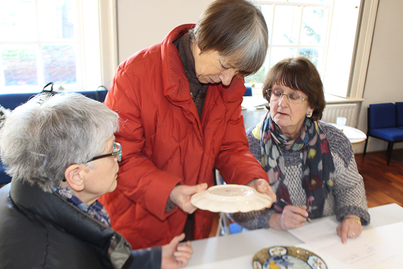 Julia looks examines the underside of a dish, with a volunteer on either side watching closely