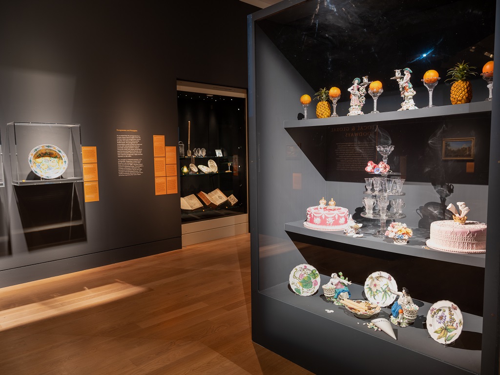 General installation view of Feast & Fast showing the low hanging height throughout, the vertically-hung pomegranate charger in its wall-mounted case, and the Confectioner’s Window display in which ‘real’ food is shown alongside the historic artefacts, which was installed partly in response to the Rowan students’ desire for food to be included to contextualize and help explain the exhibits