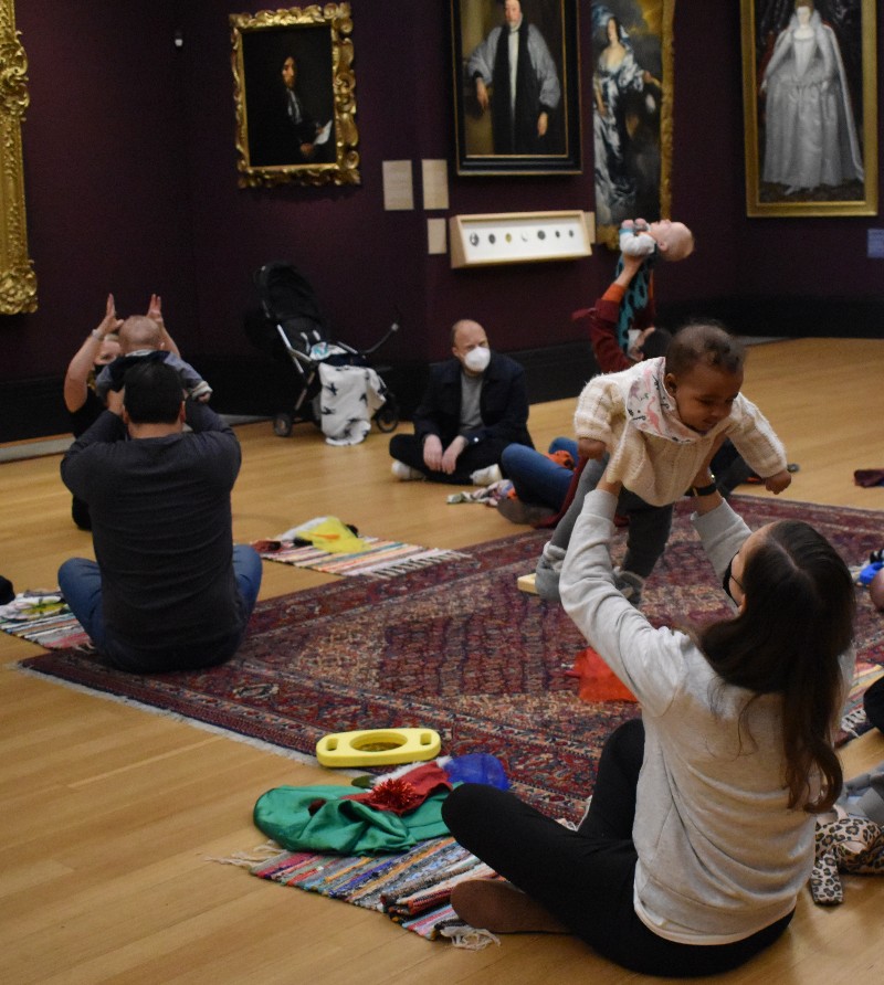 Mother lifts baby above her head while seated on a large rug in museum gallery