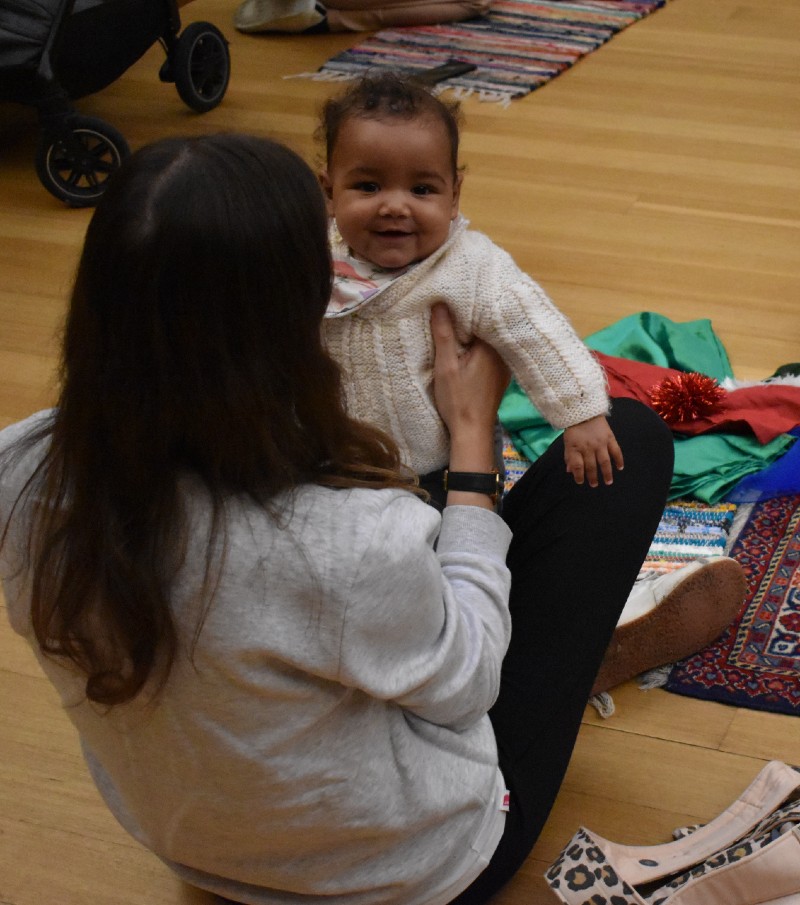 Mother sits on wooden gallery floor holding her baby who is smiling at the camera