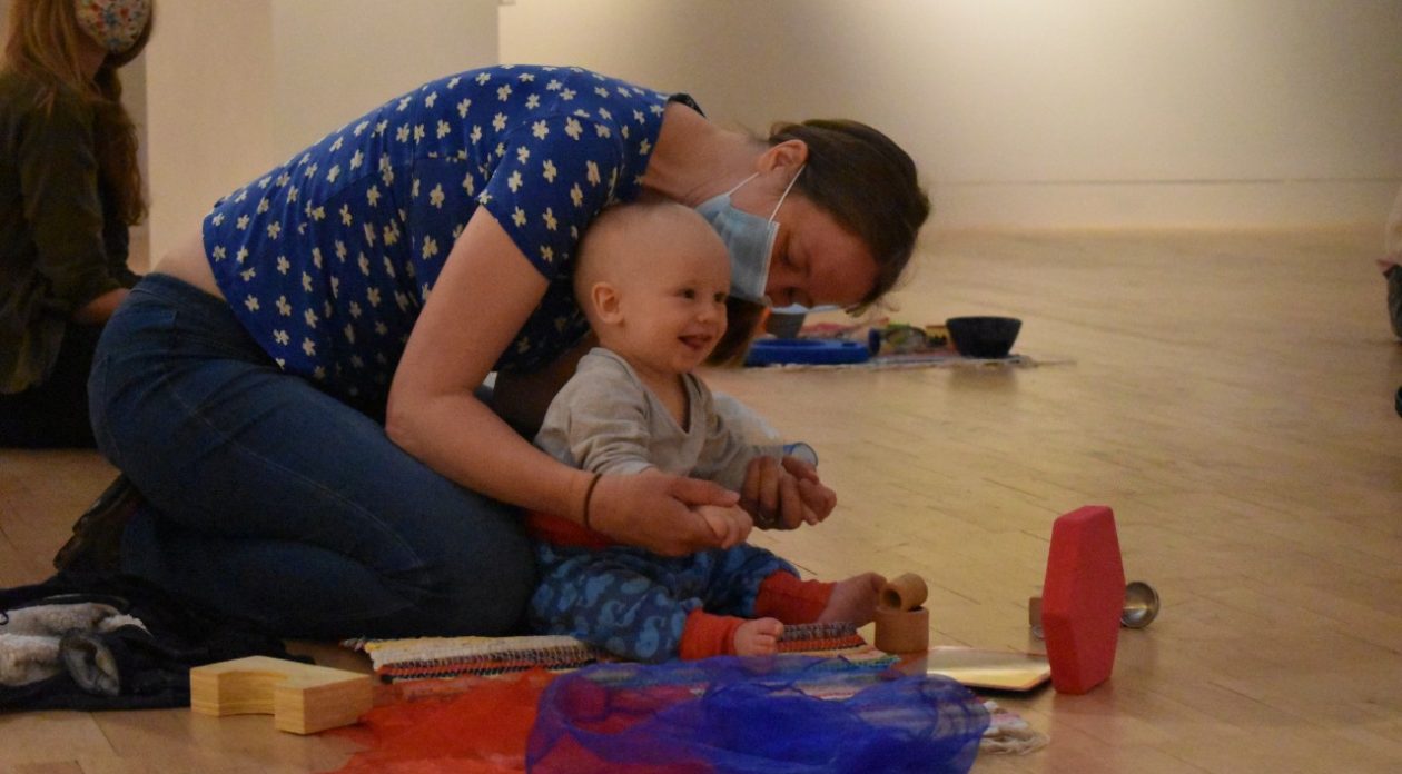 Mother kneels behind her laughing baby as they sit on wooden floor in gallery.