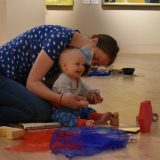 Mother kneels behind her laughing baby as they sit on wooden floor in gallery.