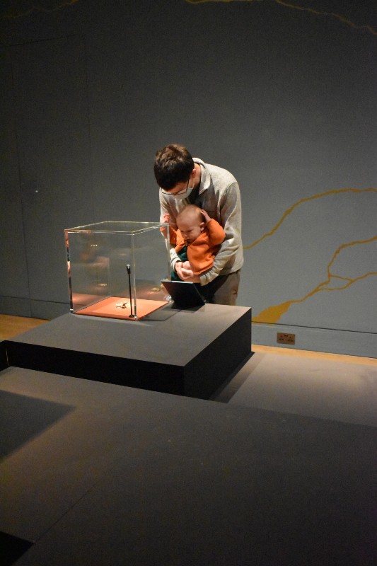 Father hold baby beside class cube display cabinet with gold exhibit 