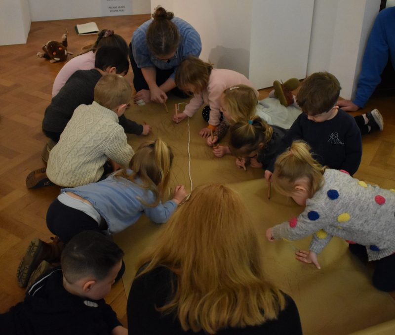 Group of 7 children and 2 adults kneel on the floor around a large piece of paper and make marks and footprints using pencils