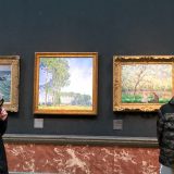A young person stands by 3 impressionist paintings hung against a dark green background