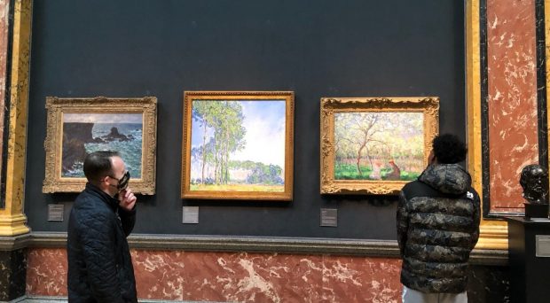 A young person stands by 3 impressionist paintings hung against a dark green background