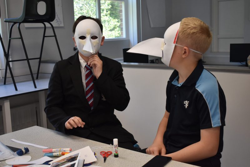 2 children sitting at a classroom table with craft materials. Both children are wearing traditional plague masks.