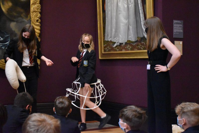 2 girls stand in front seated classmates in gallery space. One is holding a bustle, the other is wearing a cage petticoat.