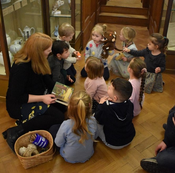 Group of small children sit on the floor in a museum gallery and listen to adult reading The Gruffalo