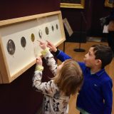 2 small children stand beside a long wall-mounted cabinet and point at the coins it displays
