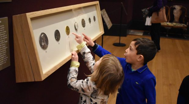 2 small children stand beside a long wall-mounted cabinet and point at the coins it displays