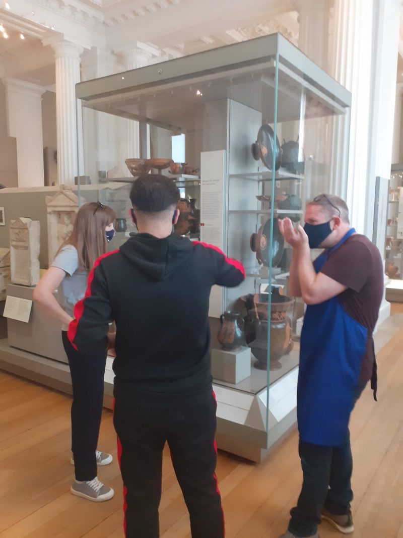 3 people stand in front of a glass cabinet displaying clay and glass vessels and amphora