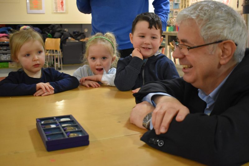 3 children sit at a table with a male adult. In front of him is a foam display case with coins