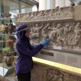 A tour participant wearing protective clear visor and disposable gloves A touch tour participant feeling the ornate carvings on an ancient Greek sarcophogus