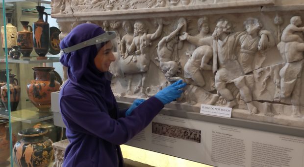 A tour participant wearing protective clear visor and disposable gloves A touch tour participant feeling the ornate carvings on an ancient Greek sarcophogus