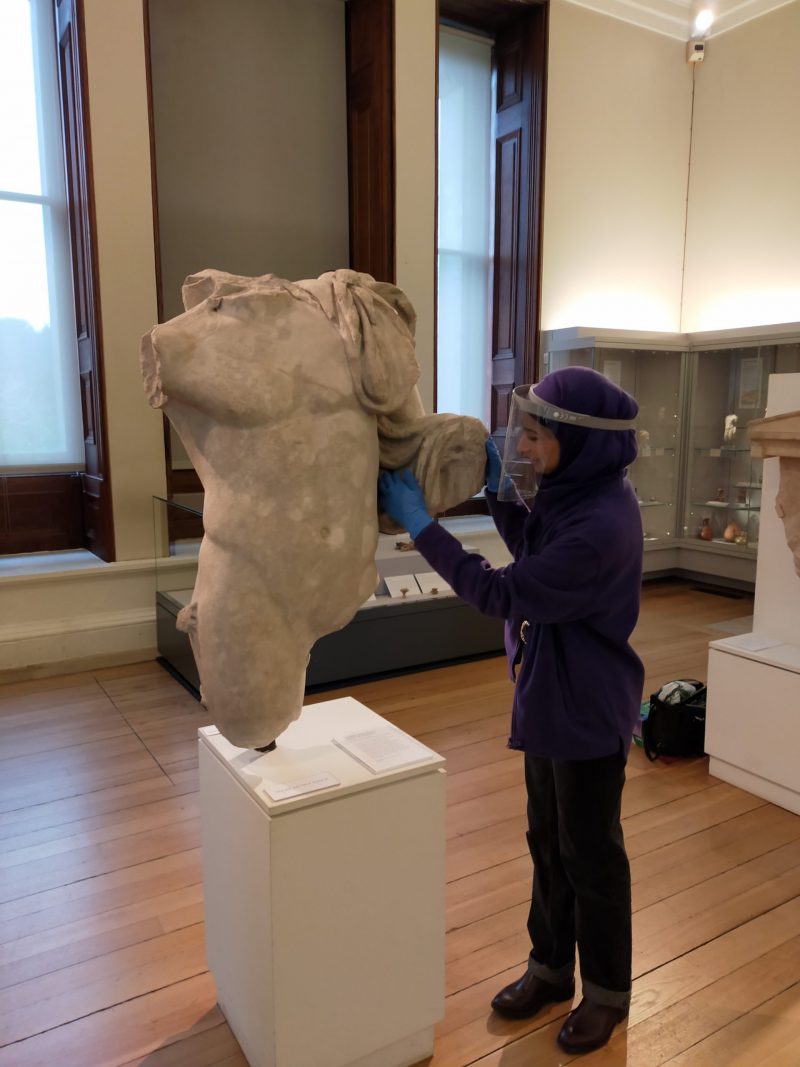 A tour participant engages with a classical Greek sculpture of a male, upper torso which is missing its arms and legs