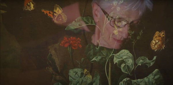 Image still from a film showing the reflection of an older woman wearing glasses in the glass of a still life with flowers painting