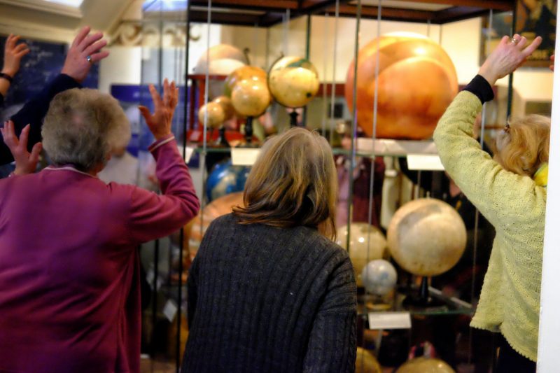 Group of older people stand round a glass cabinet filled with globes with their arms raised above their heads