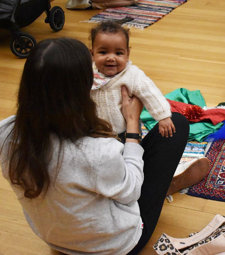 a mother holds up her smiling baby as they sit on the gallery floor with toys and activities