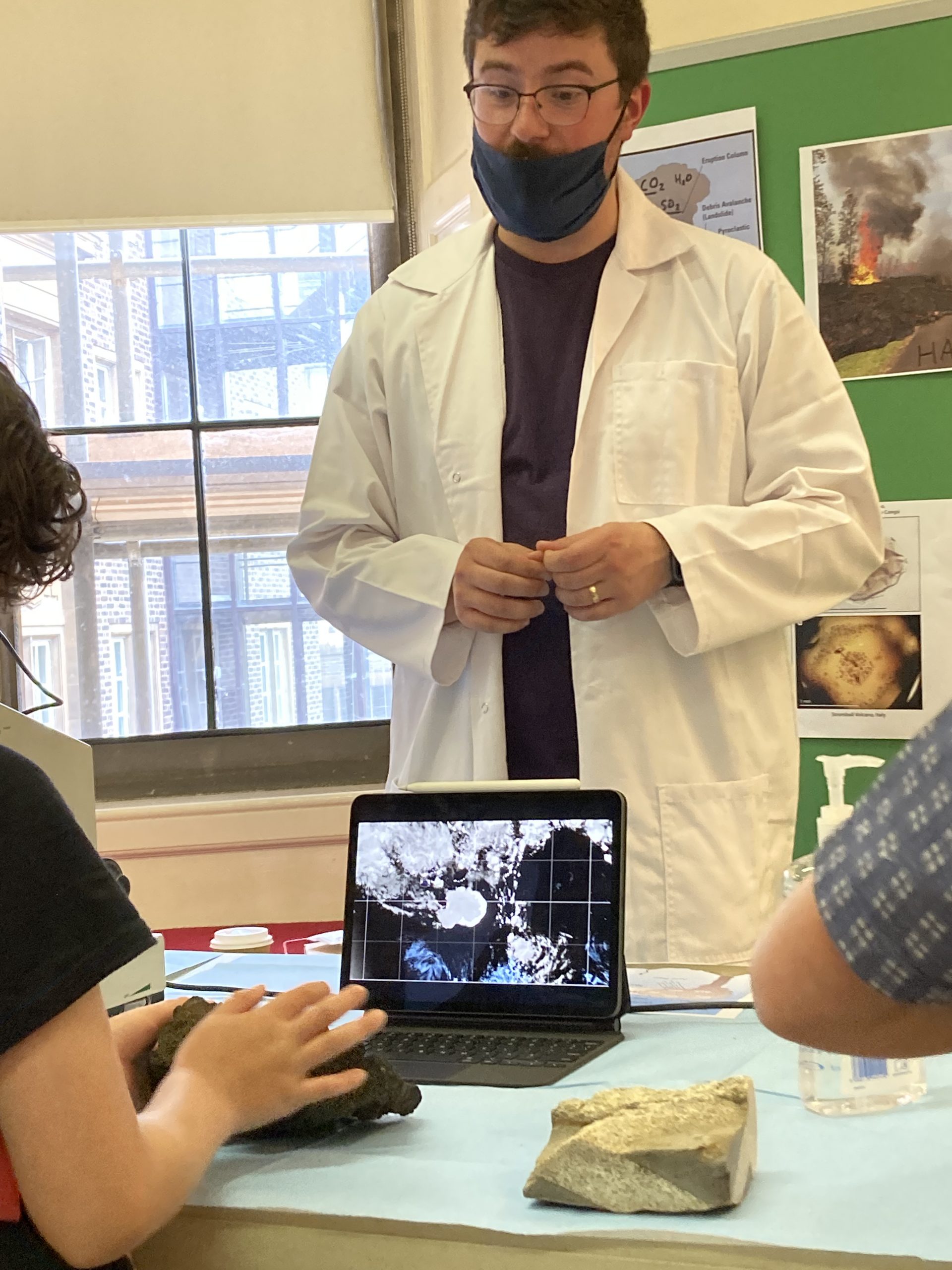 Man in white coat and mask standing behind table and speaking to visitor seated at a laptop.