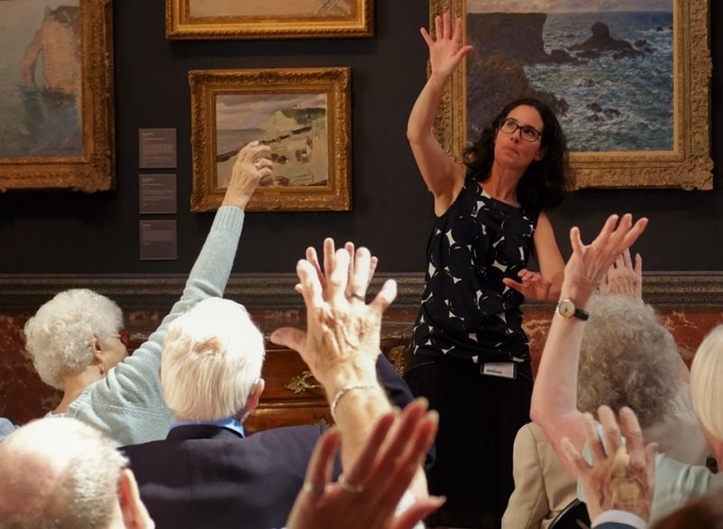 Group of seated older people raise one arm above their heads, copying the actions of a woman stood in front of them