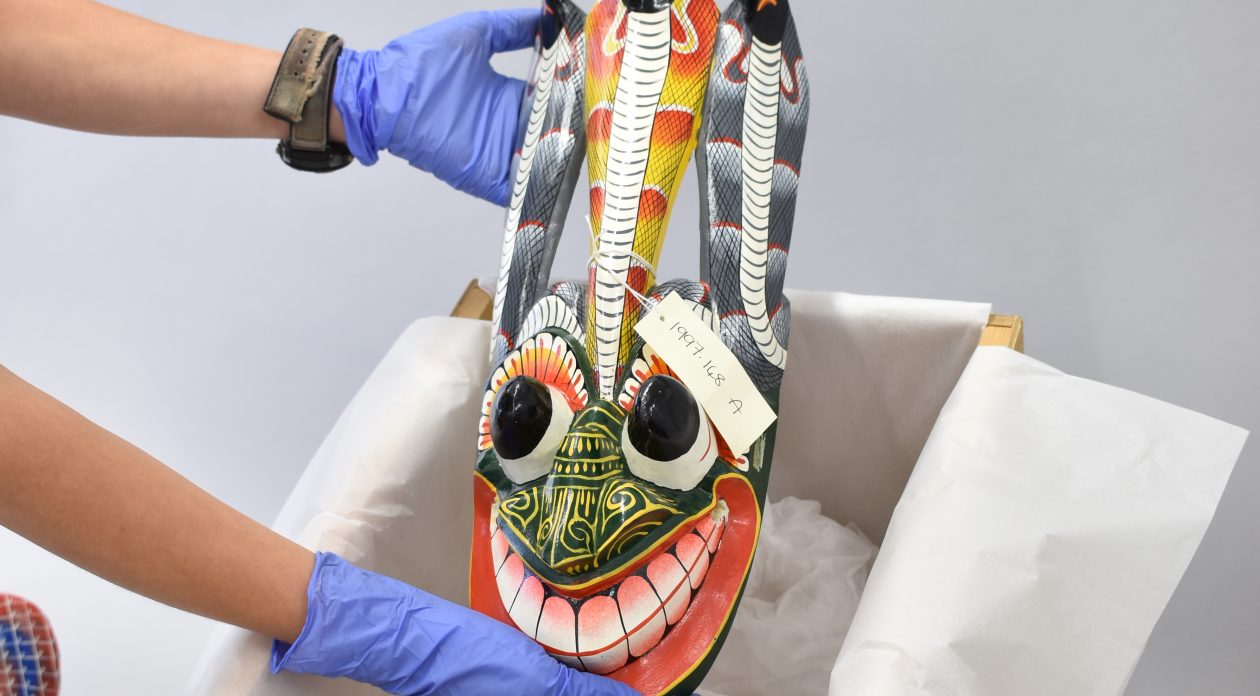 A pair of purple-gloved hands lifts a brightly coloured face-like Sri Lankan mask from its storage box.