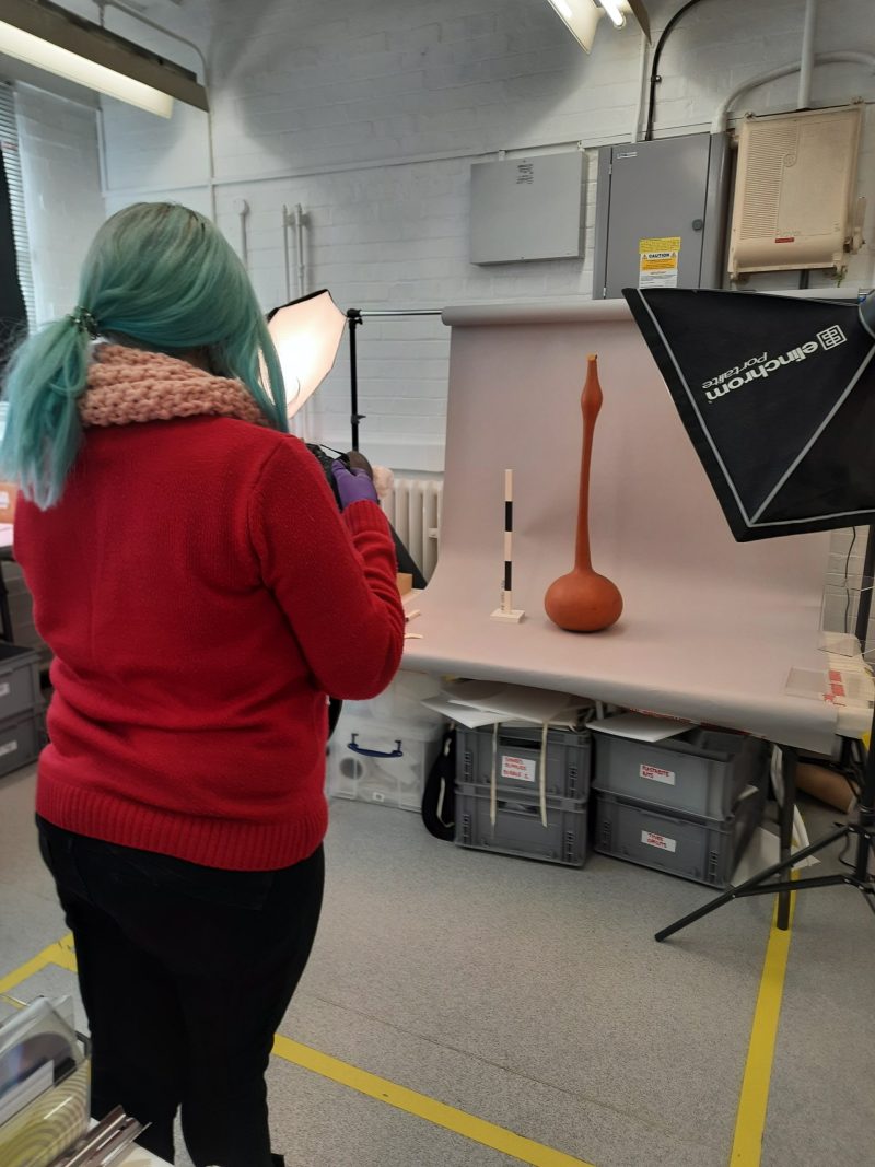 Person with long blue hair photographs a tall beer gourd against a white background
