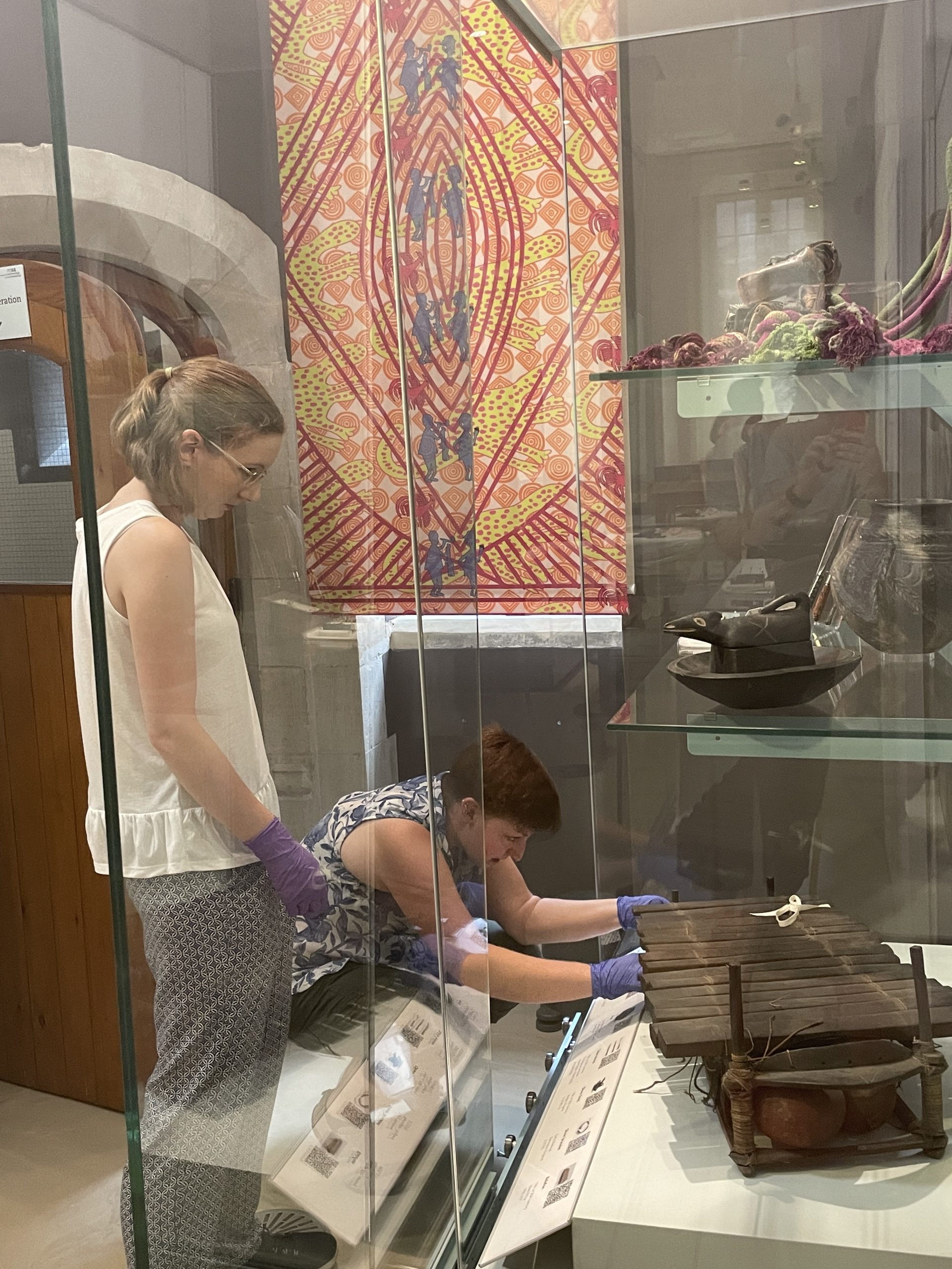 2 people (1 standing, 1 crouching) position object on to lower shelf of glass cabinet