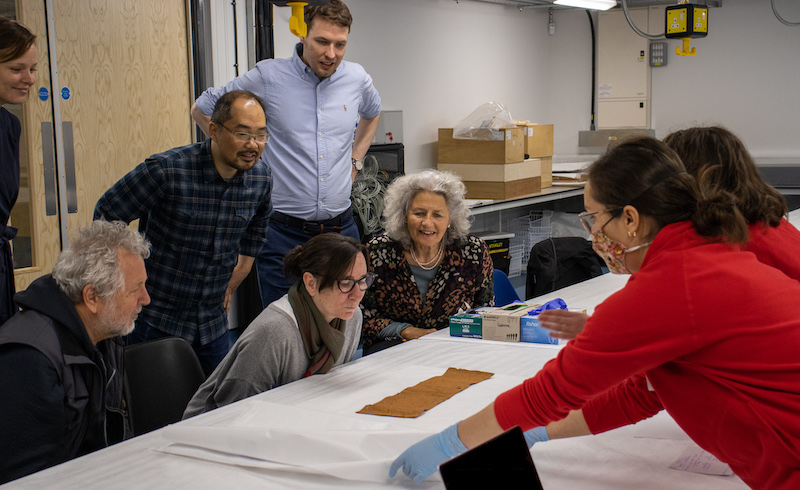 Emily and Jane presenting a handling session on the Ugandan Barkcloth at MAA Taking Care Workshop, with colleagues from Slovenia, Sweden, Germany, Austria and the UK. Image shows a small length of Ugandan Barkcloth. 3 people are sitting at a table while 5 others stand and observe. 