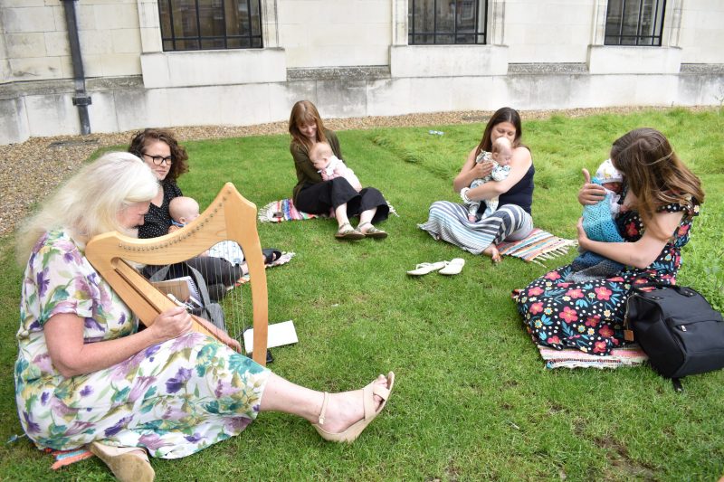4 woman and their babies sit on the grass outside the Museum. Another seated woman plays the harp.