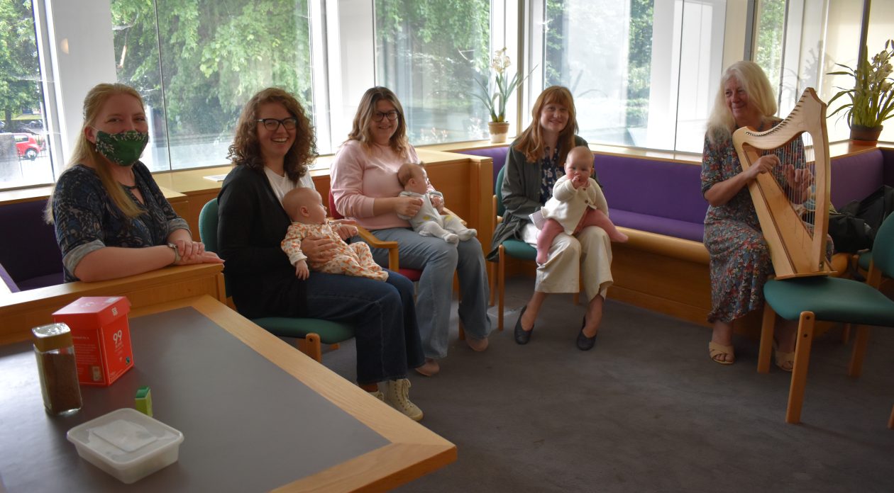 5 women sitting in a room by a window. Three have small babies on their laps and one woman is playing the harp.