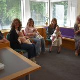5 women sitting in a room by a window. Three have small babies on their laps and one woman is playing the harp.