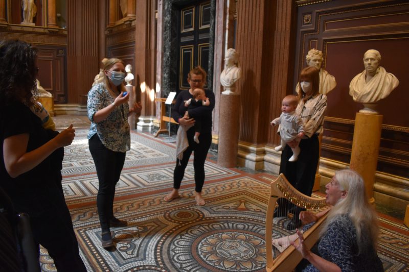 Mothers and babies in the Museum entrance hall singing while seated woman plays the harp.