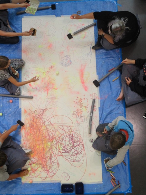Young people sitting at a long table create a group artwork on white paper using coloured powder, sticks and utensils
