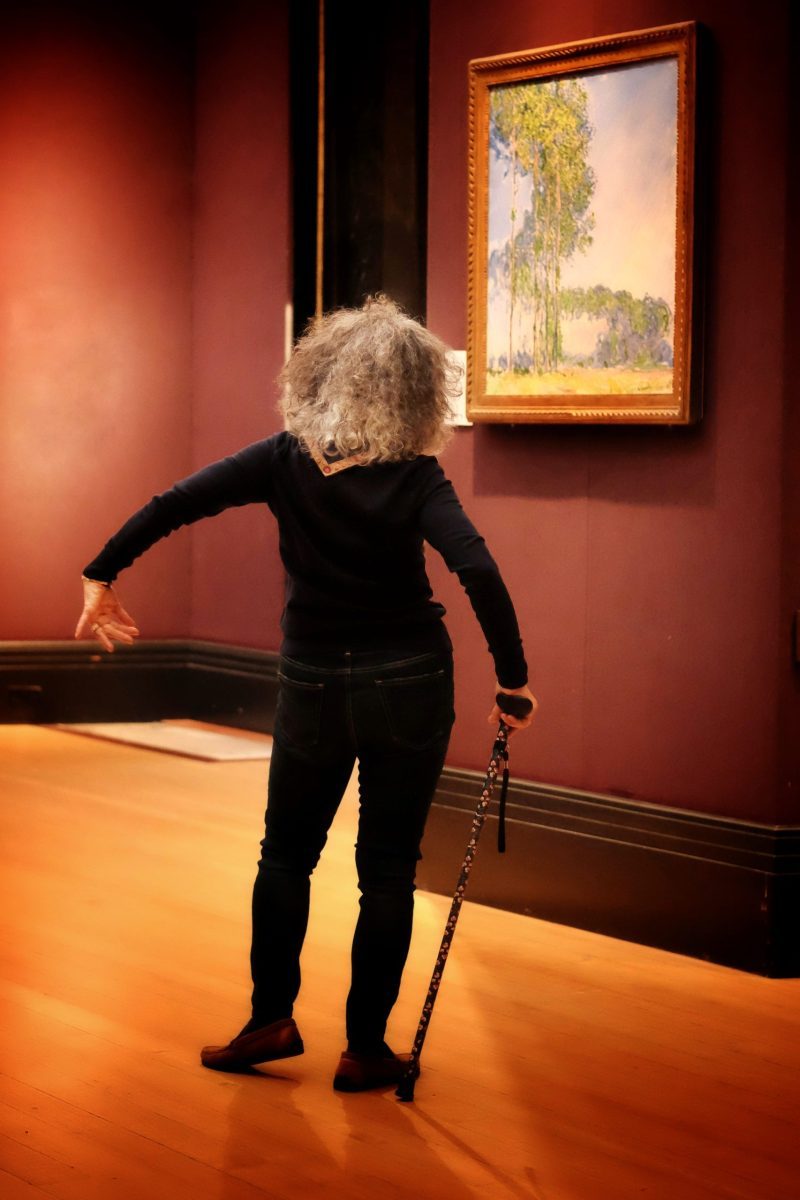 Woman with grey curly hair dressed in black stands with her back to the camera. She is holding a cane with her left arm stretched out beside her. She is standing in front of an impressionist painting of trees.