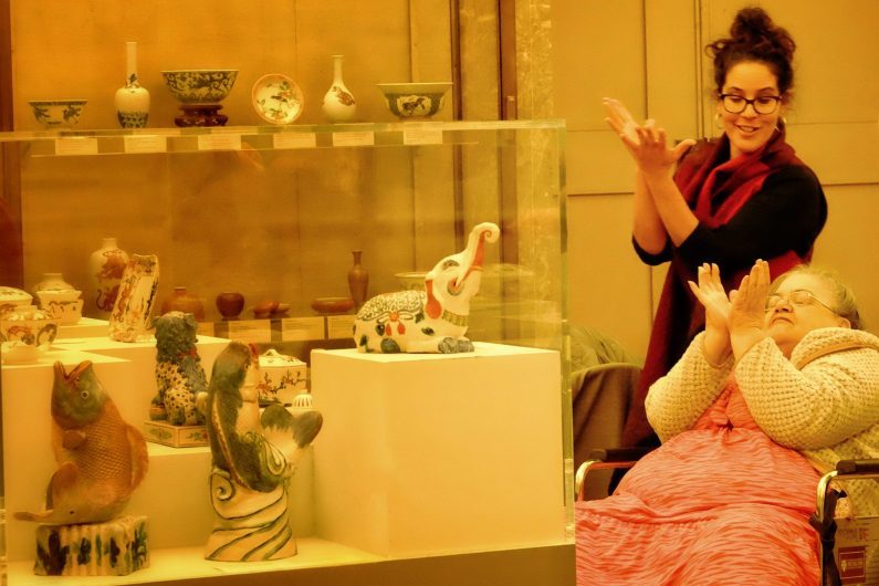 Woman seated in wheelchair in front of a glass cabinet of ceramics makes a wing-shaped sign with hands. Woman stands behind her mimicking this pose.