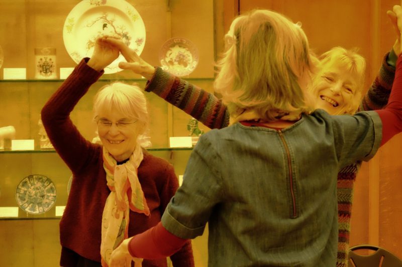 3 women link hands with arms raised. One stands with back to the camera, the other two are laughing.