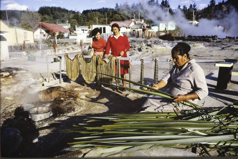 Woman seated on beach using long grasses to make a skirt while other 2 other women look on